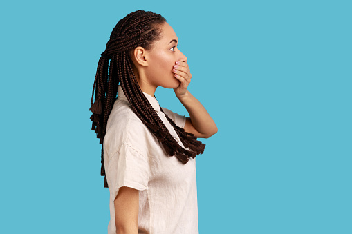 Side view of woman with dreadlocks wants to scream, covers mouth with palm, stares at something terrible, has eyes popped out, wearing white shirt. Indoor studio shot isolated on blue background.