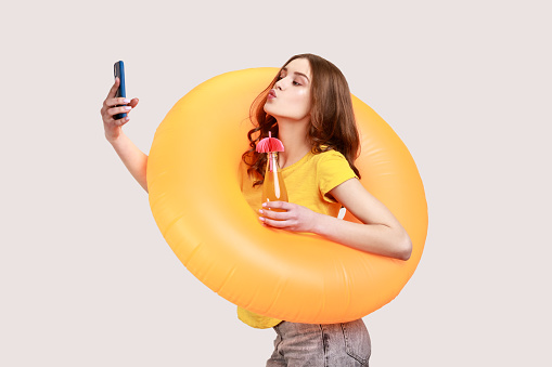 Funny female with brown wavy hair in yellow T-shirt holding orange rubber ring and making selfie at phone makes kiss gesture, having rest on resort. Indoor studio shot isolated on gray background.