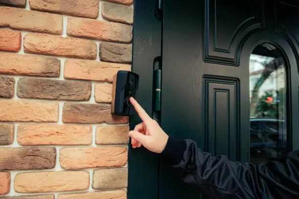 man hand in a black jacket rings the door intercom. orange brick wall