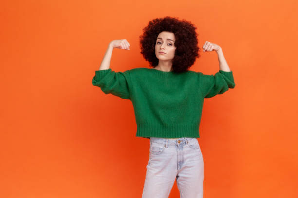 Strong confident woman with Afro hairstyle wearing green casual style sweater raising arms showing biceps, female strength and feminism. stock photo