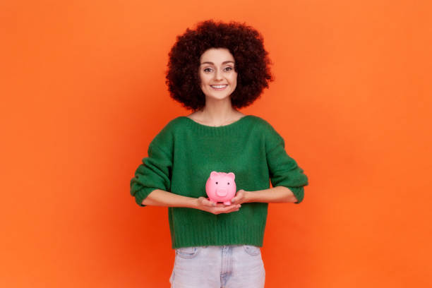 Pleasant looking woman with Afro hairstyle wearing green casual style sweater standing holding pink piggy bank in hands, saving money. Pleasant looking woman with Afro hairstyle wearing green casual style sweater standing holding pink piggy bank in hands, saving money. Indoor studio shot isolated on orange background. a penny saved stock pictures, royalty-free photos & images