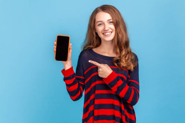 istock Woman pointing at smart phone with empty display for promotional text, looking smiling at camera. 1370691451