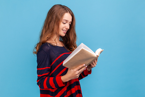 Side view of positive woman wearing striped casual style sweater, enjoying reading, standing absorbed with exciting plot, checking schedule in organizer. Indoor studio shot isolated on blue background