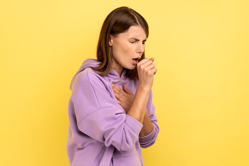 Portrait of young adult sick woman with dark hair have cough and feeling pain in her lungs, symptoms of disease, wearing purple hoodie. Indoor studio shot isolated on yellow background.
