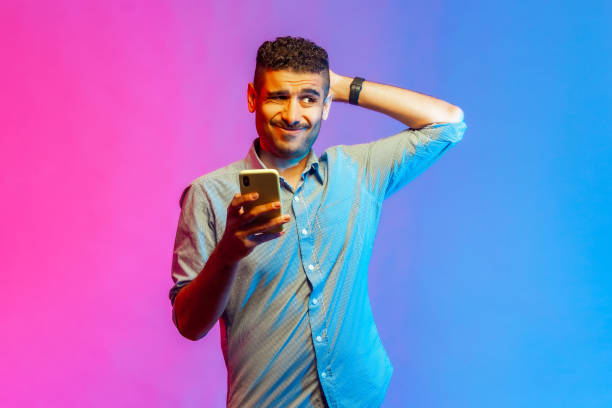 Man scratching head holding smartphone in hand, contemplating about software updating. Portrait of man in shirt scratching head holding smartphone in hand, contemplating about software updating, choosing suitable tariffs. Indoor studio shot isolated on colorful neon light background. ignoring photos stock pictures, royalty-free photos & images