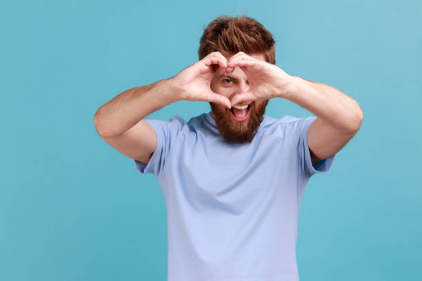 Man in blue T-shirt looking through hands in shape of heart showing romantic gesture love confession Portrait of handsome bearded man looking through hands in shape of heart showing romantic gesture, love confession, valentines day celebration. Indoor studio shot isolated on blue background. man showing peace sign stock pictures, royalty-free photos & images
