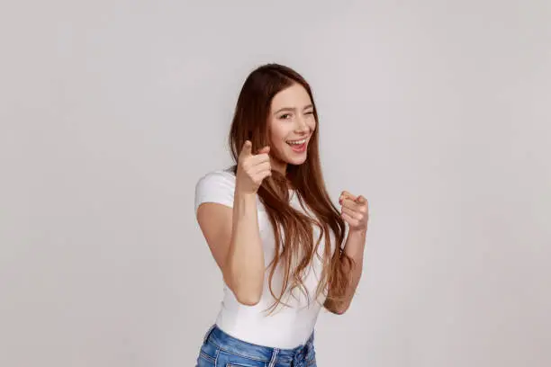 Portrait of beautiful happy woman pointing finger and winking to camera, choosing winner, gesturing we need you, wearing white T-shirt. Indoor studio shot isolated on gray background.