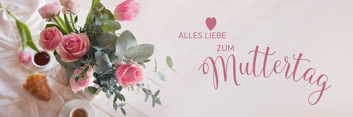 Mothers day card with pink spring flowers on white breakfast table. Horizontal top view with short depth of field and loving mother's day greeting. German text.