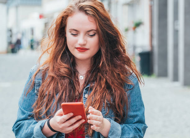 imagen conceptual de personas modernas con dispositivos tecnológicos. retrato vertical de una adolescente caucásica de cabello largo y rizado rojo caminando por la calle y navegando por internet usando el teléfono inteligente moderno. - red lipstick fotografías e imágenes de stock