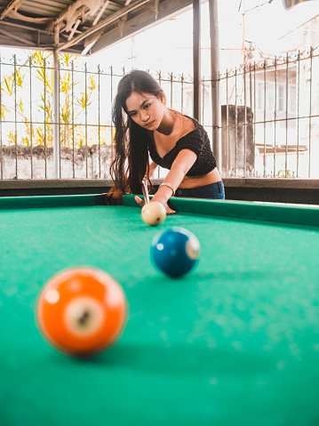 A pretty asian lady playing a game of pool aiming for the number 2 ball. At an open air and old billiard hall during the middle of the day.