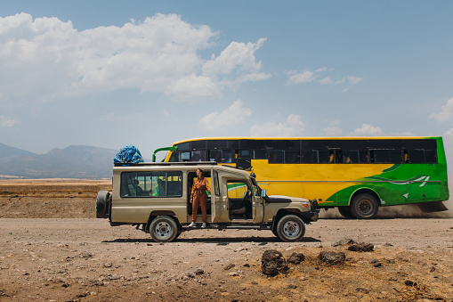 Happy woman explorer in safari jumpsuit staying by the modified jeep safari car with driving bus behind enjoying a road trip in Serengeti National park, Tanzania