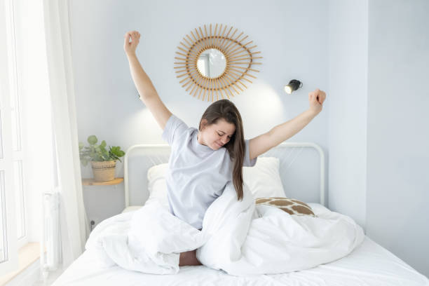 Happy cheerful girl sitting on bed after good sleep. Young woman enjoying morning on bed at home stock photo
