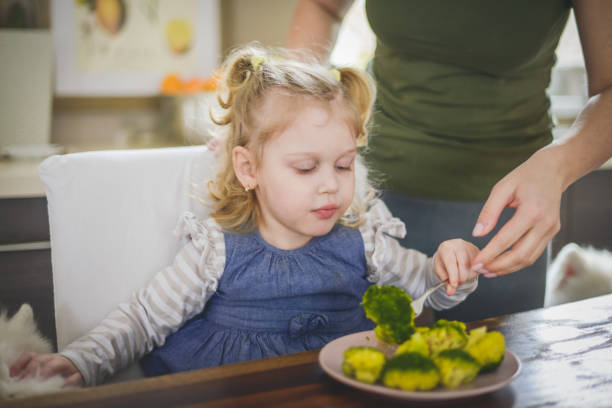 mother and baby girl blonde in home interior , eat broccoli stock photo