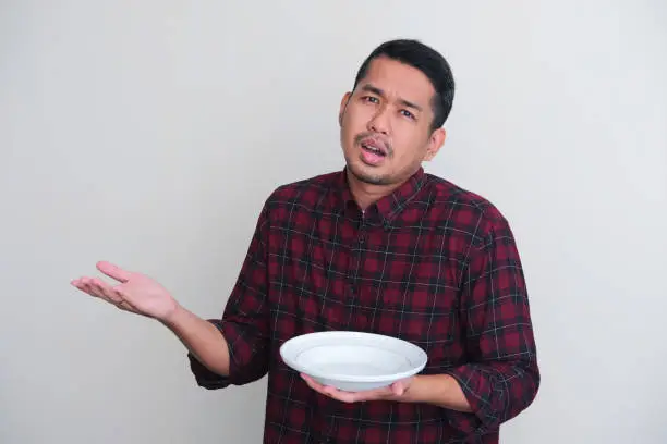 Adult Asian man showing disappointed expression while showing empty dinner plate