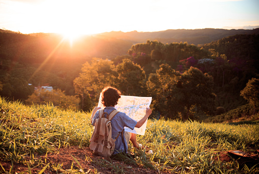 Young man looking for his location through a map, with mountains and sunset in the background. Concept of finding the right direction to achieve goals.. The map is a generic illustration made digitally exclusively for this photograph.