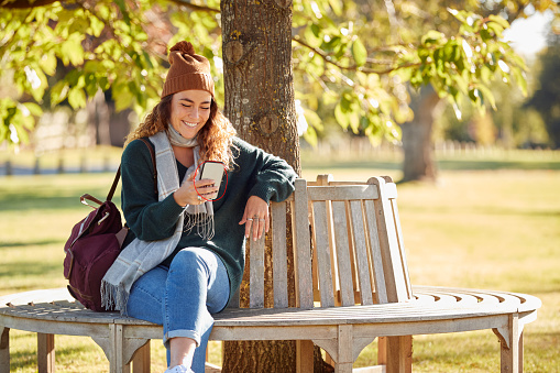 Smiling Young Woman Wearing Hat And Scarf Sitting On Bench In Autumn Park Using Mobile Phone