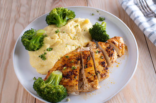 Delicious homemade cooked, gluten free meal with pan fried chicken breast, polenta and broccoli. Served on a white plate isolated on wooden table. Ready to eat