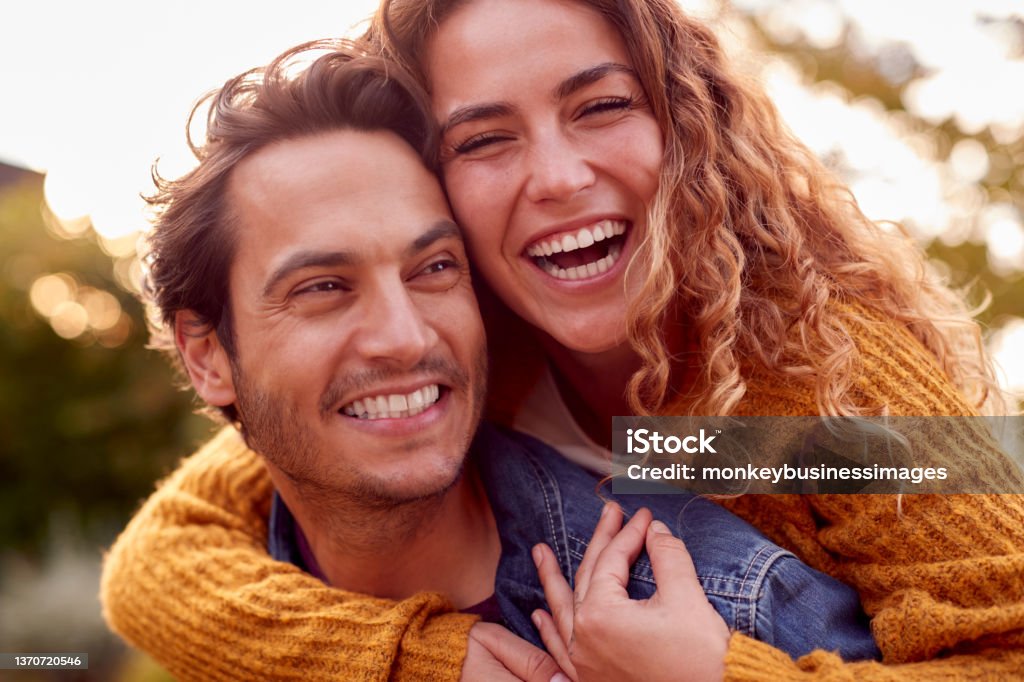 Portrait Of Happy Loving Couple With Man Giving Woman Piggyback As They Hug In Autumn Park Together Couple - Relationship Stock Photo