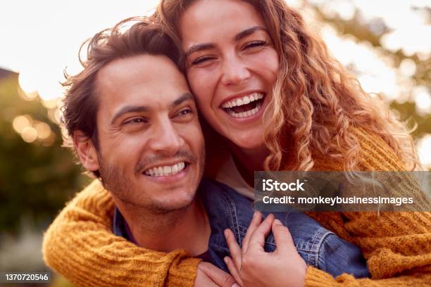 Portrait Of Happy Loving Couple With Man Giving Woman Piggyback As They Hug In Autumn Park Together Stock Photo - Download Image Now