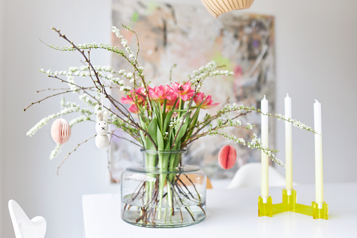 A modern Easter bouquet with tulips and twigs stands in a bright environment on a white table in a glass vase