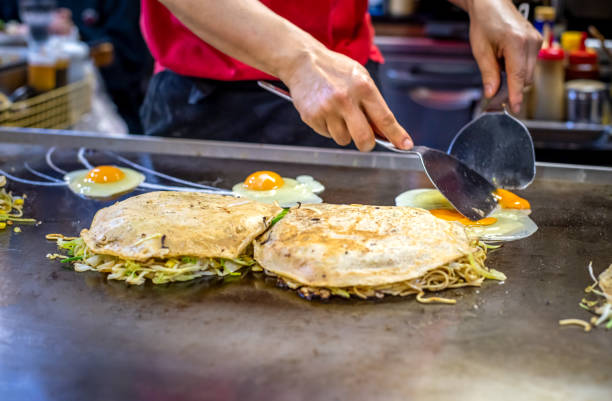 hef is cooking Okonomiyaki with eggs chef is cooking Okonomiyaki with fried eggs on the iron plate, he is using the spatula to put the food on to the plate which is already done. Typical from Osaka and Hiroshima kinki region stock pictures, royalty-free photos & images