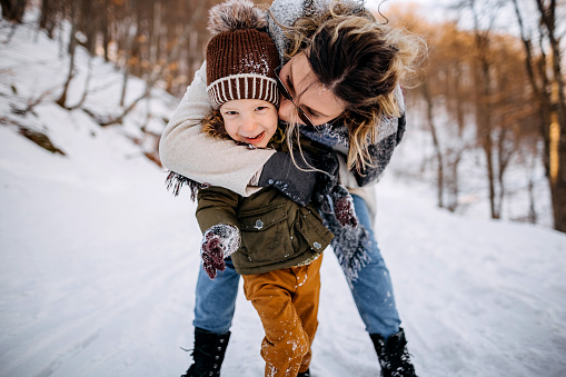 Young happy mother enjoys with her little son in a beautiful mountain winter idyll. They walk and play in the snow.