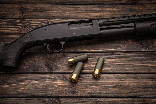 A black 12-gauge pump-action shotgun and ammo are on a wooden table. A smooth-bore weapon with a plastic stock. Dark brown background.