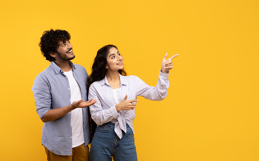 Excited indian couple cheerfully pointing aside at empty space, yellow studio background, panorama. Young hindu man and woman aiming at advertisement, copy space