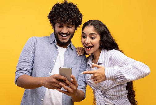 Portrait of excited indian couple using cellphone and pointing on it, looking at phone with open mouth, posing over yellow background, banner