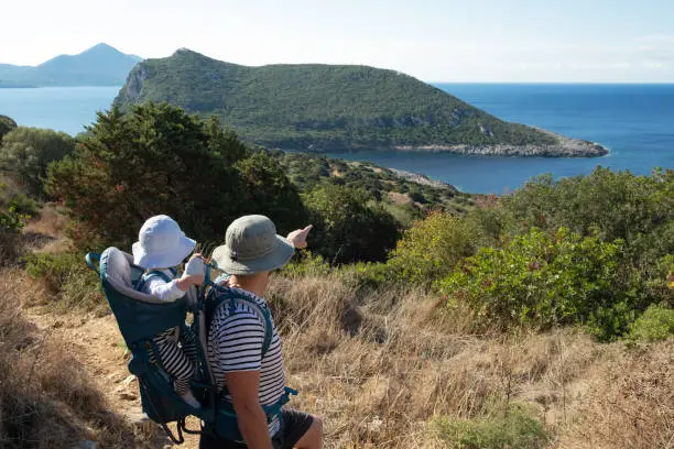 Photo of Caucasian man carrying backpack with baby wearing sunhat, travelling in beautiful places in nature in summer sunny weather,observing breathtaking views pointing at mountains,sea.Family hiking,treking
