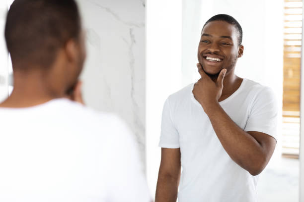 istock Happy Young Black Man Looking In Mirror In Bathroom And Touching Face 1370731314
