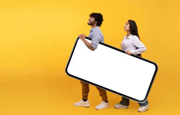 Photo of Young indian couple holding giant smartphone with mockup for design, yellow studio background, free space