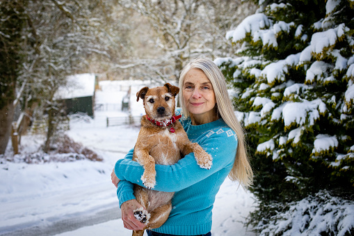 A portrait of a mature woman wearing a sweater, holding her senior patterdale terrier while standing outdoors in the snowy Northumberland weather. They are both looking at the camera and the woman is smiling.