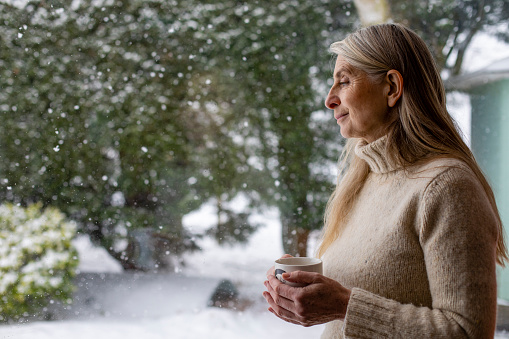 A mature woman wearing a warm sweater while standing indoors against a window in her house and having a hot drink in a mug. She is looking outdoors at the snowy scene outdoors.