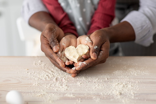 Hands of black father and daughter holding heart shaped cookies together, unrecognizable african american dad and child baking pastry at home, enjoying time together, closeup, cropped shot