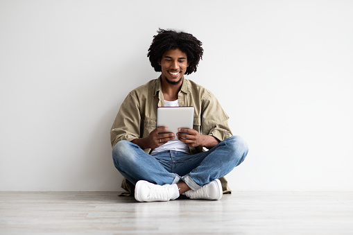 Cheerful Millennial Black Guy With Digital Tablet Relaxing On Floor Indoors, Happy Young African American Man Sitting Near White Wall At Home And Using Modern Gadget For Browsing Internet, Copy Space