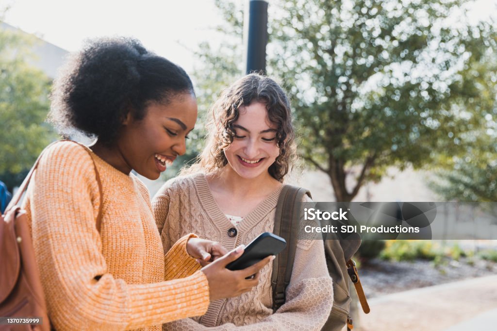Teen girl shows friend something on her smart phone The teenage girl shows her female friend something on her smart phone that makes both of them smile. Friendship Stock Photo