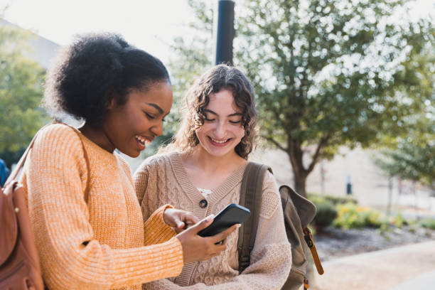 Teen girl shows friend something on her smart phone The teenage girl shows her female friend something on her smart phone that makes both of them smile. adolescents hanging out stock pictures, royalty-free photos & images