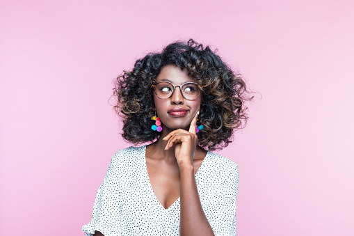 Pensive african young woman wearing white dress, colorful earrings and eyeglasses, looking away with hand on chin. Studio portrait on pink background.