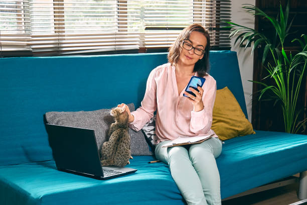 istock Woman with glasses sitting on sofa with her cat and using smartphone checking email news online, looking for friends in internet social networks, writing blog or made call by work. Freelance working. 1370742586
