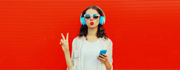Portrait of stylish young woman posing in headphones listening to music with smartphone on red background stock photo
