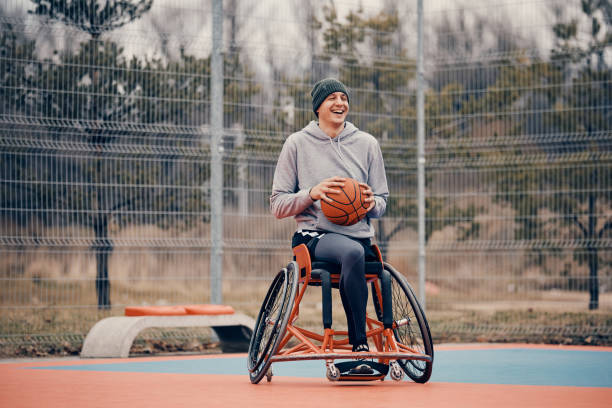 Cheerful amputee in wheelchair having fun while playing basketball on outdoor court. Happy basketball player with disability uses wheelchair while playing on outdoor sports court. adaptive athlete stock pictures, royalty-free photos & images