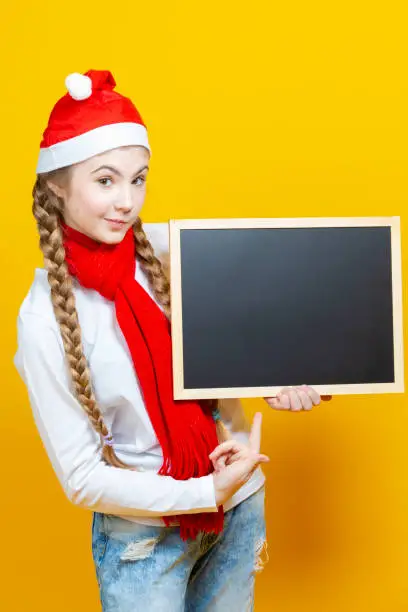 One Caucasian Girl In Santa Hat and Red Scarf Posing With Black Wooden Blackboard While Pointing With Pointfinger On Yellow Background. Vertical Image