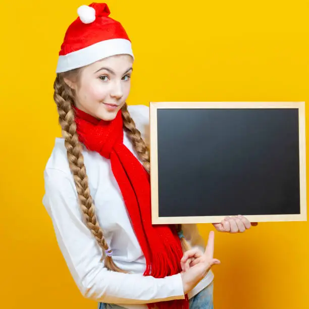 Intriguing Caucasian Girl In Santa Hat and Red Scarf Posing With Black Wooden Blackboard While Pointing With Pointfinger On Yellow Background. Square Image