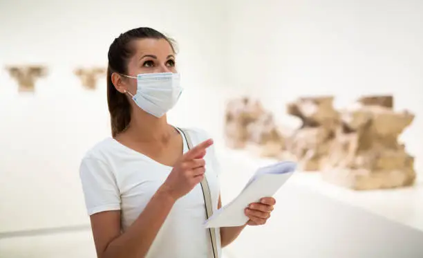 Portrait of young woman in protective face mask observing sculptures exposition in art museum, local tourism during pandemic situation