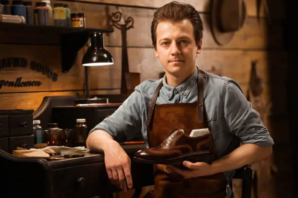 Photo of young cheerful shoemaker in workshop holding shoes. Looking at camera.