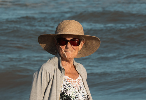 an older woman looking up at the camera smiling. she has a hat and sunglasses. In the background you can see the sea with its beautiful blue color and its waves.