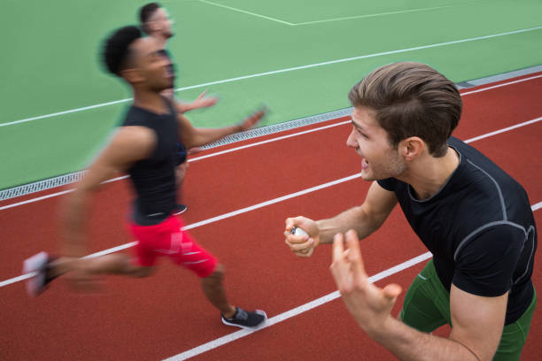 A small group of adults are seen hitting the finish line together as they complete a race.  They are each dressed comfortably and are smiling with their arms raised, as they break through the red ribbon with excitement.