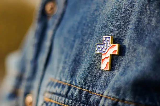 Photo of Close up of Christian cross pin with American flag colors is pinned on blue jeans jacket. Patriotism and religious rights concept