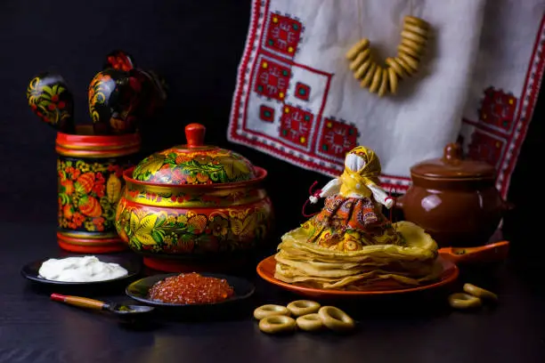 Traditional Russian food and symbols on dark background for Maslenitsa festival. Pancakes, caviar, sour cream, rushnik, textile doll and wooden utensils with Khokhloma painting for Shrovetide.